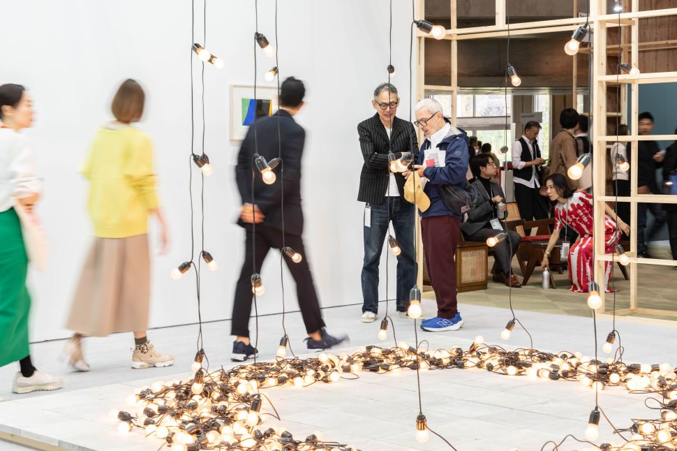 Visitors walk through a fair booth installation with strings of hanging lightbulbs encircling a glowing arrangement on the floor; some people stop to talk while others move past the illuminated display.