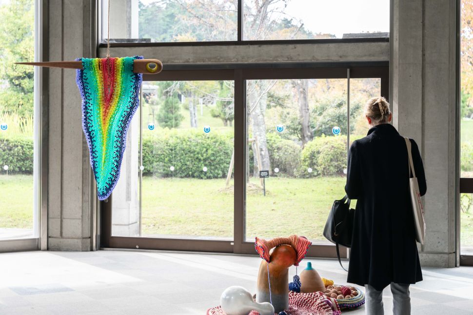 A visitor stands in front of a windowed gallery space, viewing a colorful textile and mixed-media installation that includes a rainbow-hued hanging piece resembling fabric on a spindle and sculptural forms arranged on the floor.