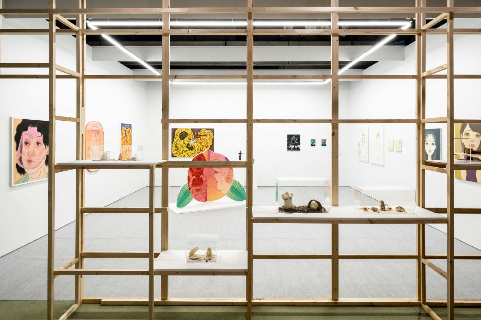 View of an exhibition booth framed by a wooden lattice structure, showing a variety of colorful contemporary paintings and small sculptural works. Portrait-style paintings with exaggerated features hang on white walls, while vitrines display small torso sculptures and organic forms on shelves and plinths.
