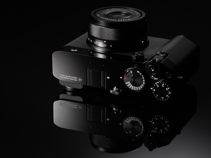A black digital camera with several control dials and a textured lens is shown on a glossy, reflective surface, with "Fujifilm X-Pro3 Fragment Edition" labeled on the top.