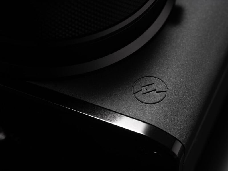 Close-up of a sleek, black electronic device featuring a circular surface and a raised logo with two slashes inside a circle, captured in dramatic lighting with metallic highlights.