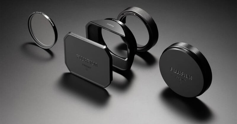 Five Fujifilm camera lens accessories, including caps, filters, and a lens hood, are displayed in a row on a dark surface with dramatic lighting, highlighting their metallic and matte finishes.