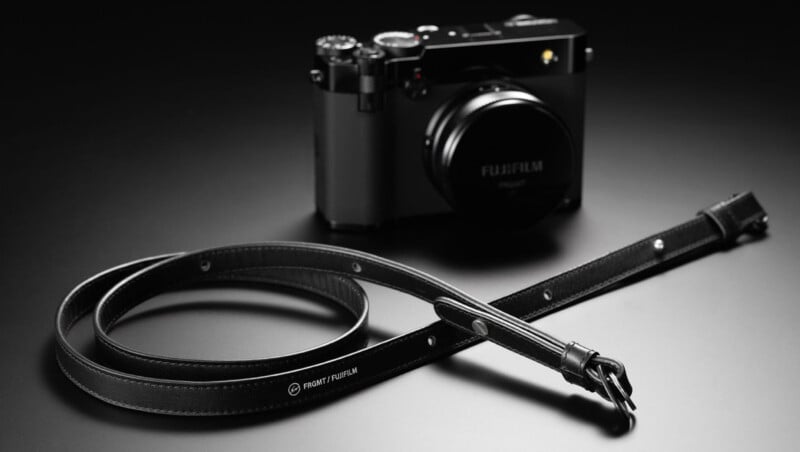 A black Fujifilm digital camera sits on a dark surface with a coiled black leather camera strap, labeled "Front Fujifilm," placed in the foreground. The camera and strap are lit by soft, dramatic lighting.