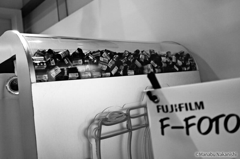 A container filled with many rolls of 35mm film sits on a counter in a store. A partially visible FUJIFILM F-FOTO sign is in the foreground. The image is in black and white.