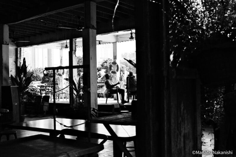Black and white photo of a covered outdoor patio with tables and chairs. Two people sit and talk in the background, framed by pillars, with plants and city buildings visible beyond.