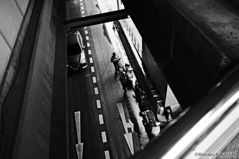 Black and white photo taken from above shows a person standing next to parked scooters on the side of a road, with a car passing by and large structures casting shadows overhead.