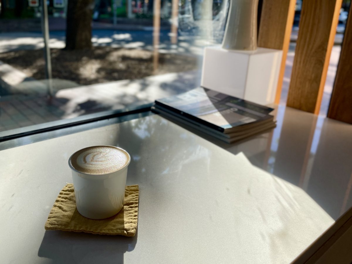 A coffee in simple white cup sits on a square of yellow fabric as the sun slants through the window.