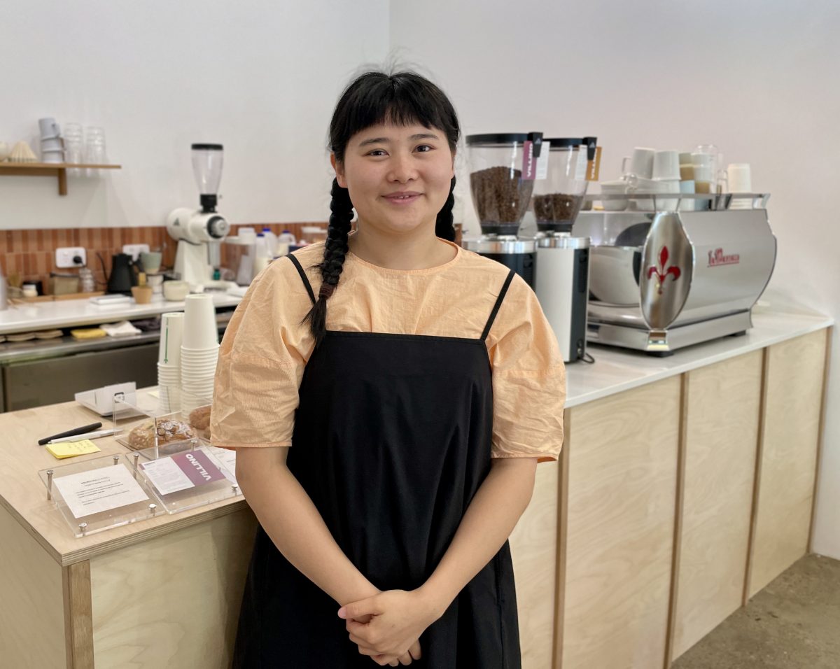 A woman with black hair in two plaits and a fringe wears a black apron over light orange blouse. She stands in front of a cafe counter. 