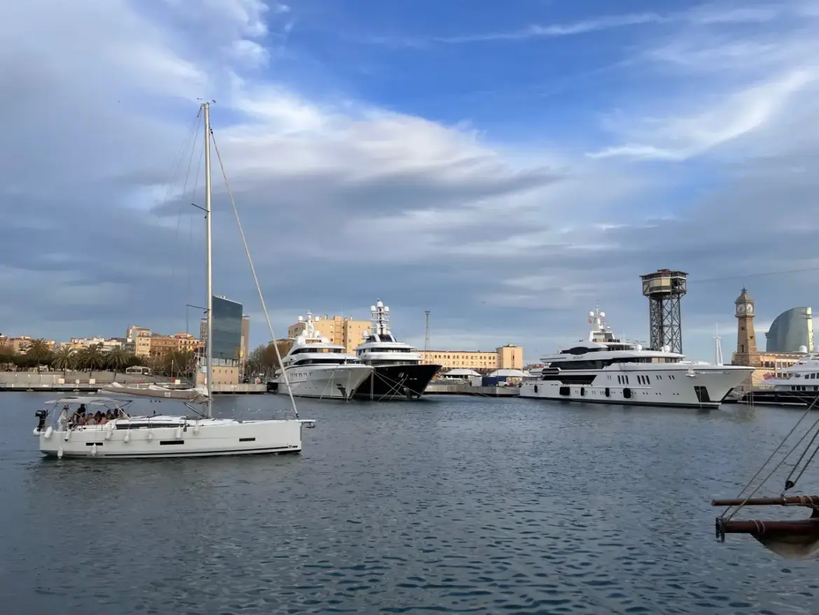Luxury yachts docked at Barcelona’s Marina Port Vell with a sailboat passing by and city landmarks in the background.