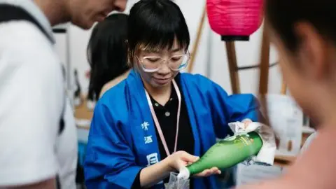 HyperJapan A bespectacled Japanese woman with short black hair wearing a blue kimono with white lettering pours sake rice wine from a green bottle into a glass.