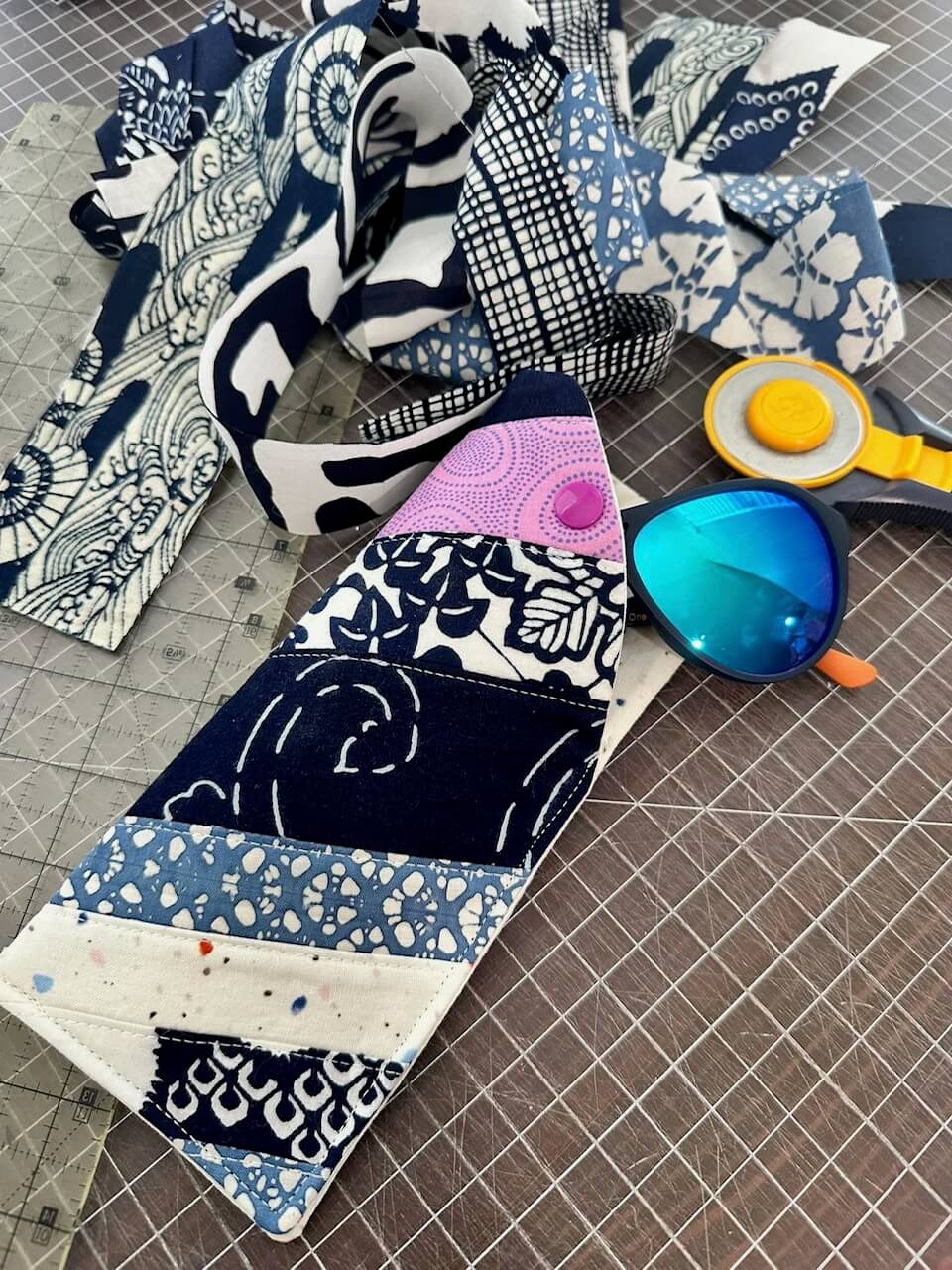 Swaths of various fabric and fabric rotary cutter lay on a fabric mat next to a sewn sunglasses holder holding a pair of plastic sunglasses in Navy spouse's business, Mottainai Creations.