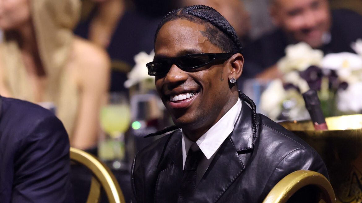 Everything You Need to Kno… Travis Scott in a black leather jacket and sunglasses is smiling at an event, with people and flowers blurred in the background.