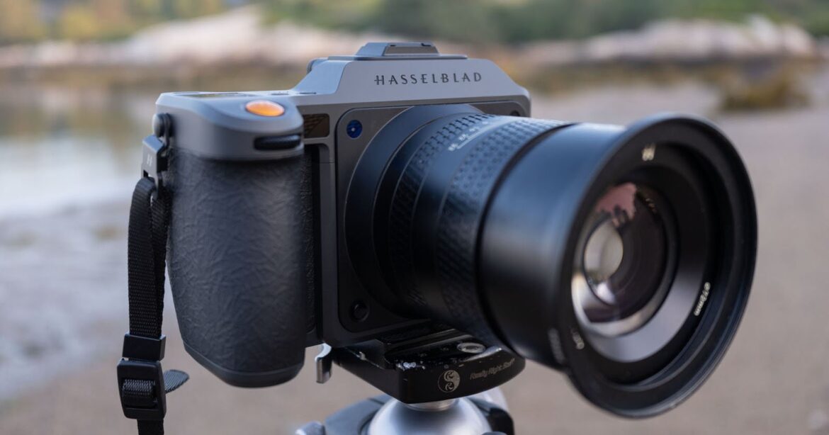 A close-up of a Hasselblad camera mounted on a tripod outdoors, with a blurred natural background. The camera’s lens and textured grip are clearly visible.
