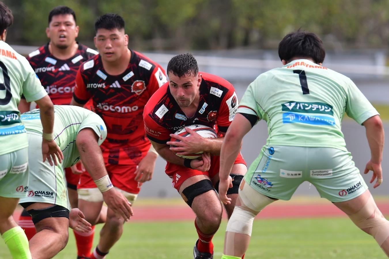 A rugby player in a red and black jersey carrying the ball, surrounded by players in light green jerseys.