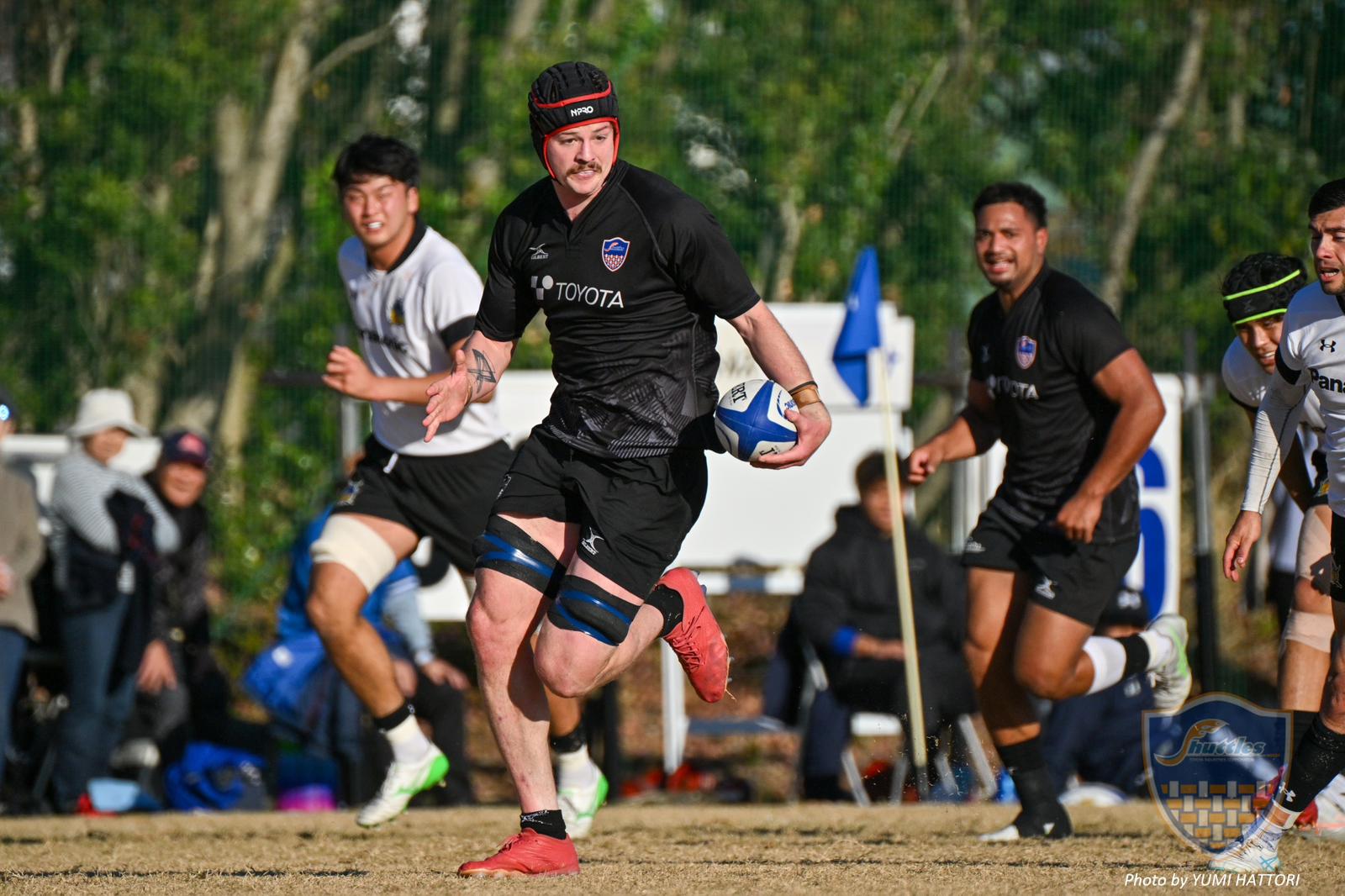 A rugby player in a black jersey with "TOYOTA" written on it is running with the ball on a field during a game.