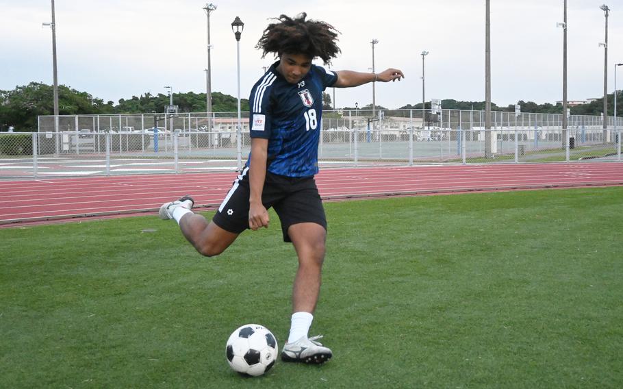 A teenager wearing a blue jersey and black shorts kicks a soccer ball on a field, with a track and a chain link fence in the background.