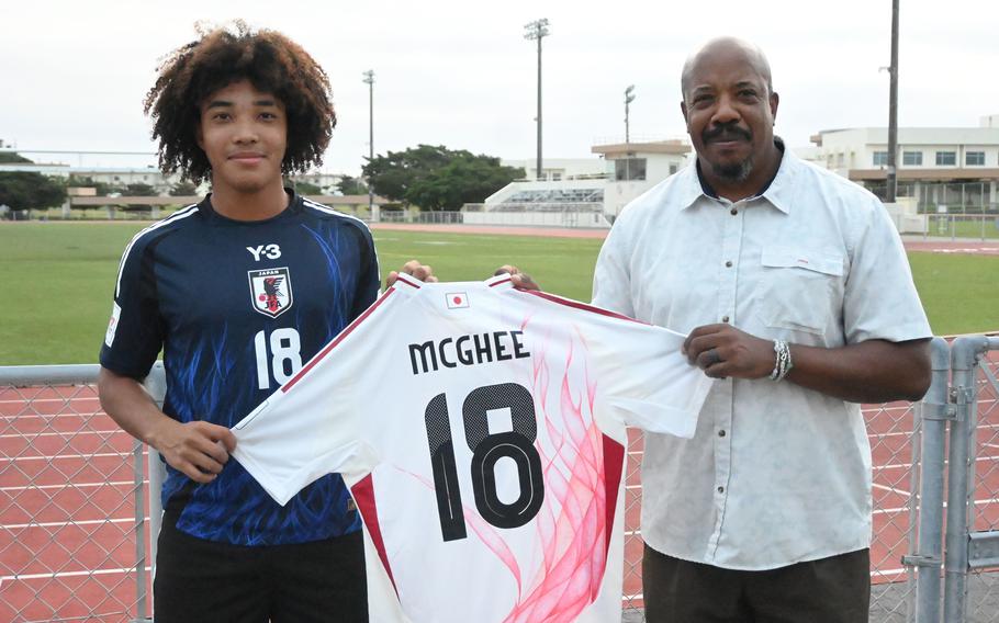 A teenager and his father pose while holding up a white No. 18 soccer jersey with the name McGhee between them.