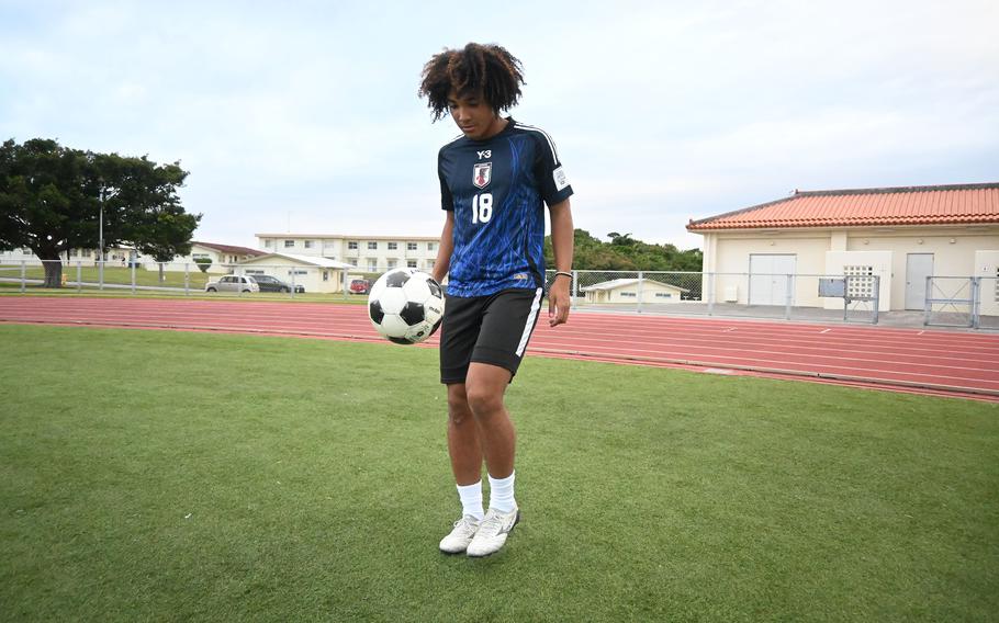 A teenager wearing a blue jersey and black shorts dribbles a soccer ball on a field with a track and school buildings in the background.