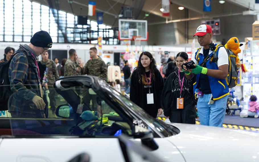 Two young women in black hoodies observe a man in a blue jacket taking a photo of a man in flannel shirt and black beanie posing in front of a convertible car.