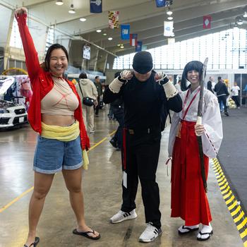 Three Comic Con attendees pose on the convention floor: a woman wearing blue shorts and a red jacket; a man wearing a black shirt, black pants and black hat; and a woman wearing a white and red kimono and holding a sword.