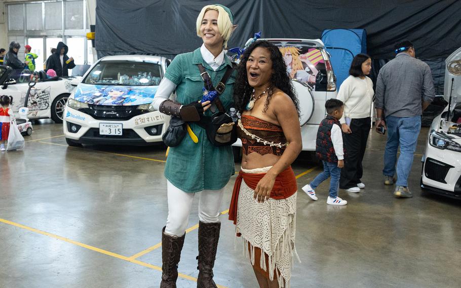 Two Comic Con attendees pose on the convention floor with two cars in the background: a man in a green jumper, white pants and borwn boots; and a woman in a brown and orange Hawaiian islander-style crop top and white skirt.