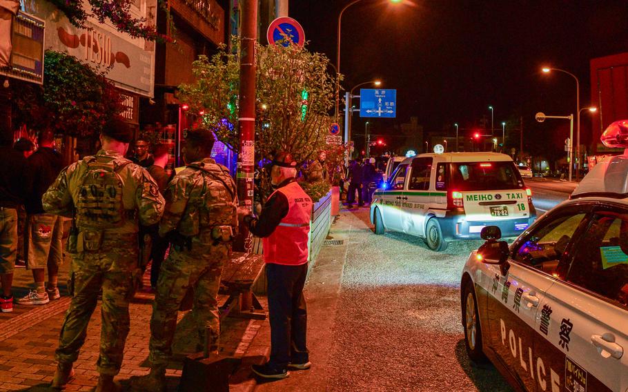 Two military members in camouflage uniforms stand on a city sidewalk with a civilian police officer in an orange reflective vest. A police car is parked in the street next to the sidewalk, and the night air is illuminated by street lamps.