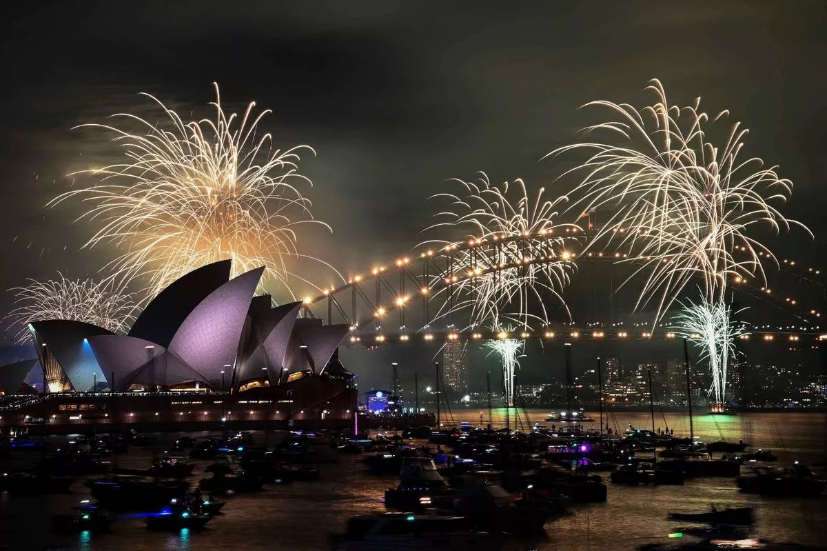 Fireworks light up the night sky over a famous architectural landmark, with numerous boats on the water and spectators gathered nearby.