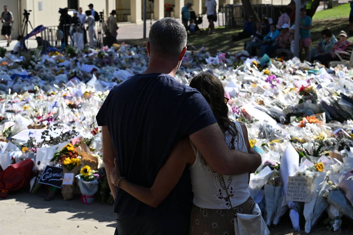 Two individuals are standing close to one another, facing a vast display of flowers and tributes set up outdoors, with a backdrop of a building and a gathering of people.