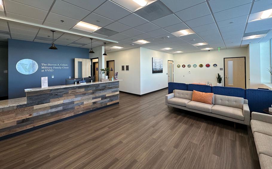 The waiting area of a medical office is seen, with a brown wooden floor, a couch on the right of the frame and a reception desk on the left.