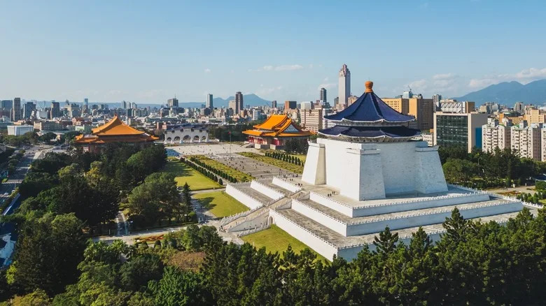 Aerial view of Taipei's Chiang Kai-shek Memorial Hall with its blue-tiled roof, flanked by the National Theater and National Concert Hall, set against the modern city skyline and distant mountains