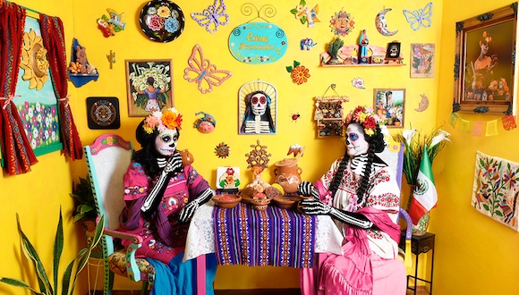 2025 LaSelle Foundation Fellows to Travel to Austria & Japan Two women in full Catrina costume sit at a table with cafecito and pan, the yellow walls decorated with many colorful folk art objects.