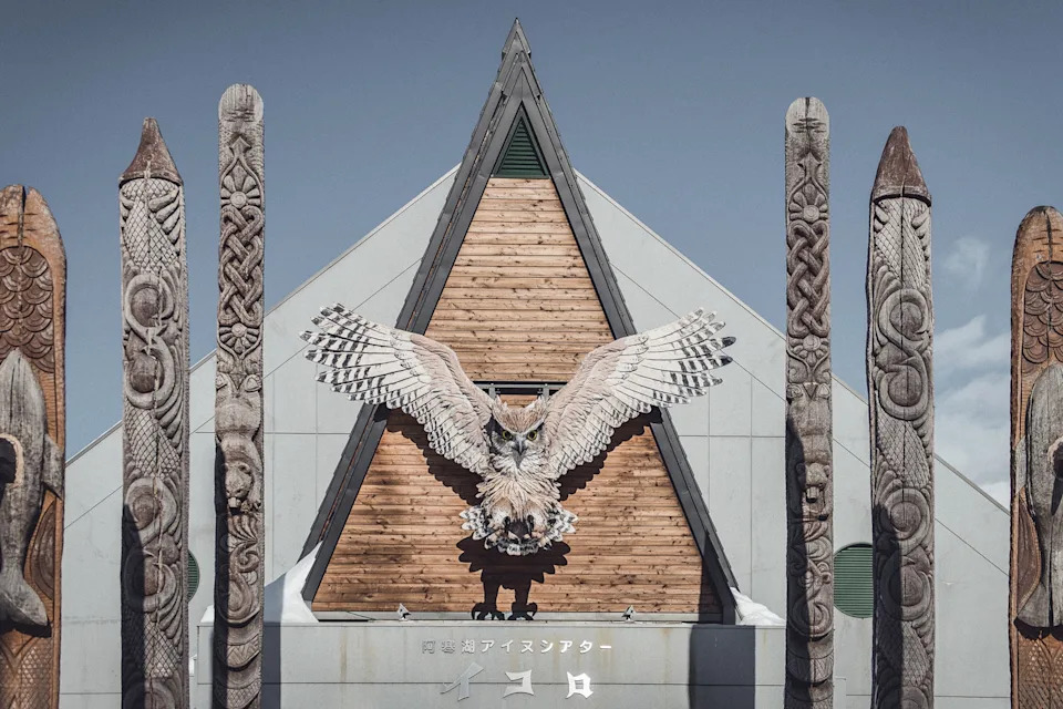 The wooden facade of a tent-like building with carved totem poles on either side of the entrance and a carved, wooden owl towering above.