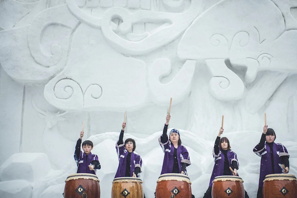 A group of indigenous, Japanese women at different ages, as well as a boy, pounding on big barrel drums in front of a sculpted ice wall.