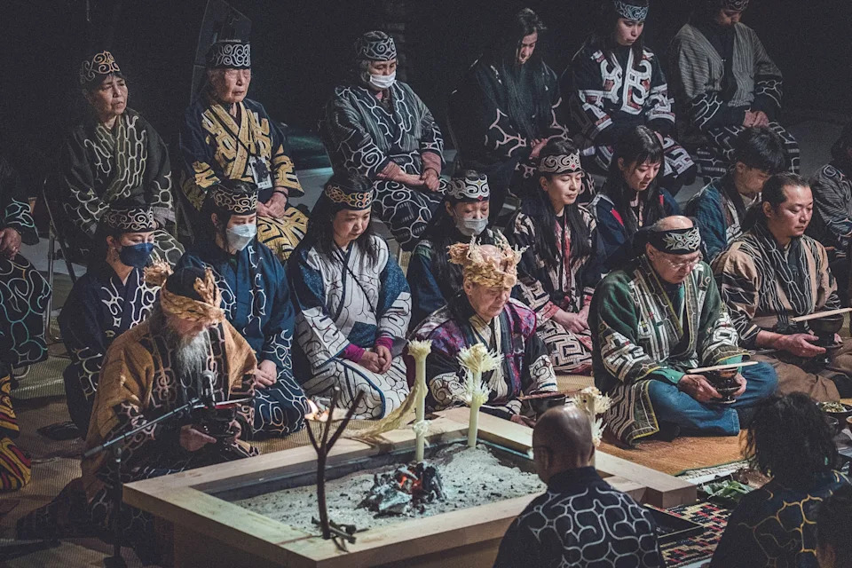 The moody and dark interiors of a ceremonial room with rows of indigenous Ainu people sitting cross-legged on the floor in traditional clothing.