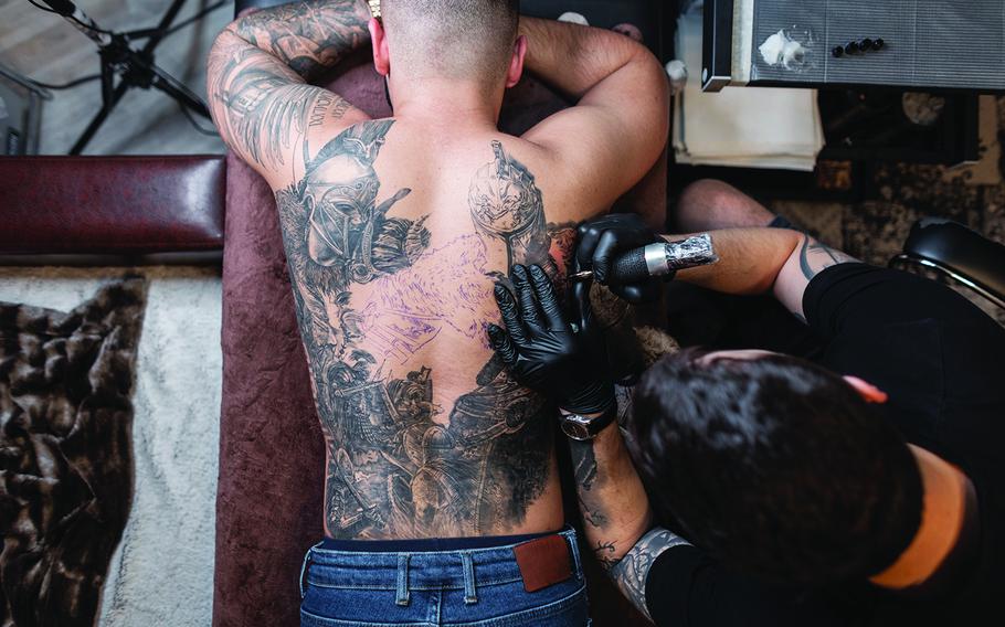 Tattoo artist man with black gloves draws a knight tattoo on the guy’s back in the studio, top view. Tattoo artist’s workflow