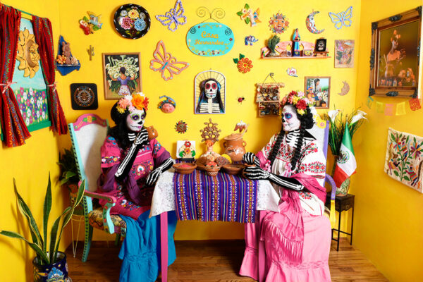 Two women in full Catrina costume sit at a table with cafecito and pan, the yellow walls decorated with many colorful folk art objects.