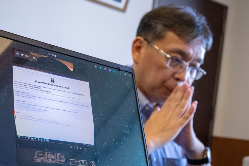 A laptop displaying a screenshot of a ransomware demand, with a man seated next to it.