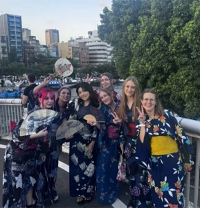A group of young women wear Japanese kimonos. They stand on a bridge over a river and display fans and make peace signs with their fingers.