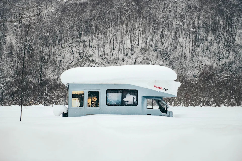 A campervan bus buried deep under a layer of powdery snow in a winter forest landscape.