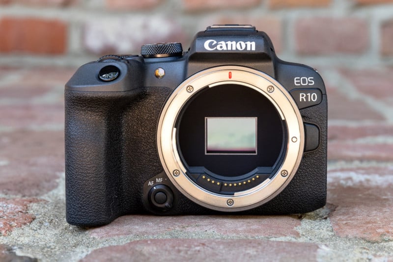 A Canon EOS R10 mirrorless camera body is shown without a lens attached, revealing the sensor and lens mount, placed on a brick surface with a blurred brick background.