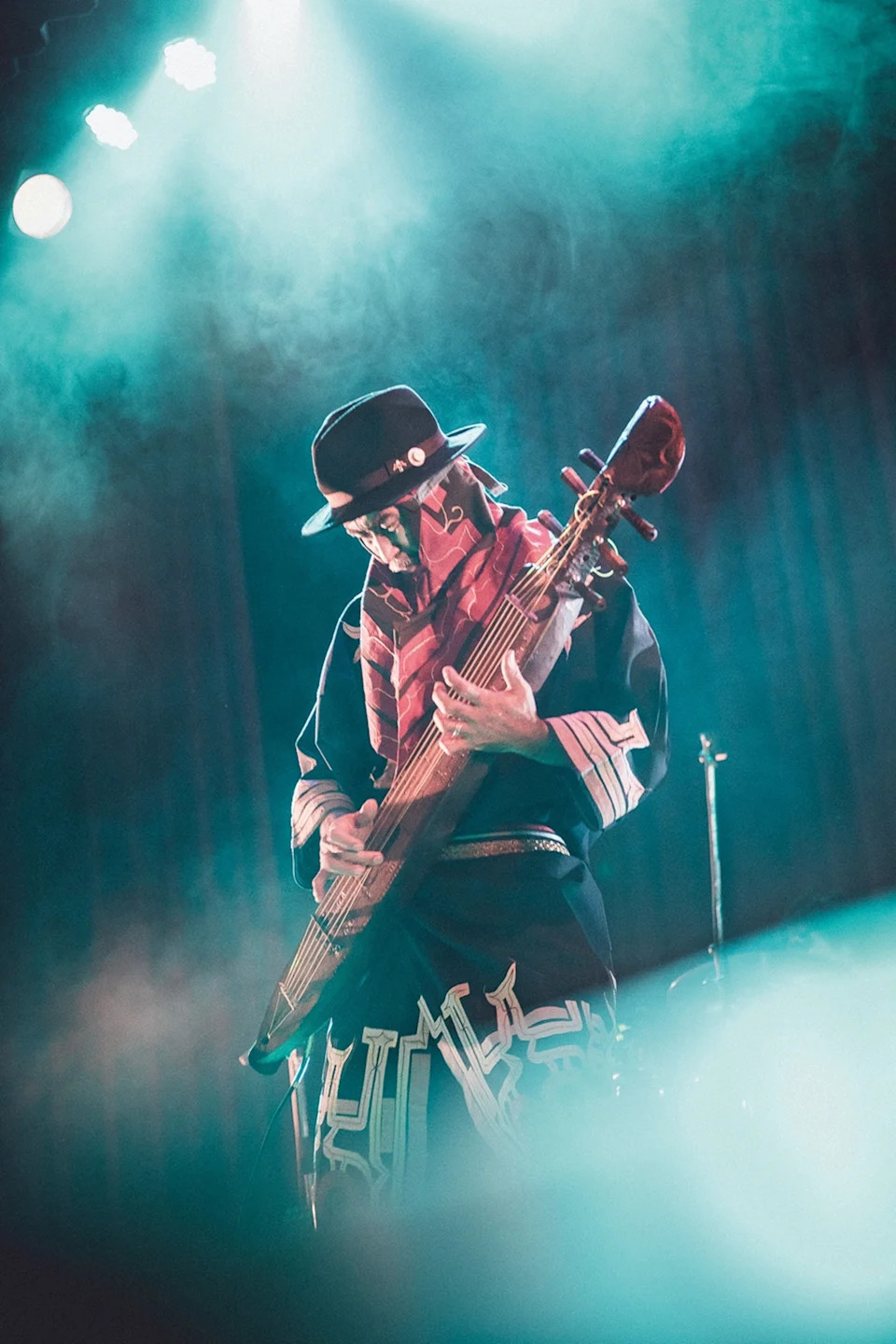 A dynamic concert shot of an eldery man in fedora on stage, playing a traditional, guitar-like harp.
