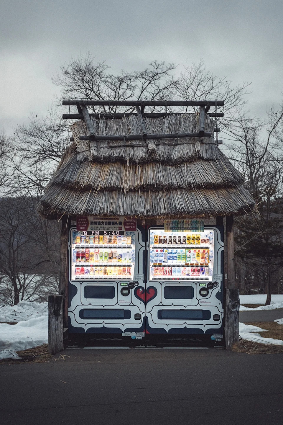 A snowed-in vending machine with a straw roof on the side of a road at late dusk.