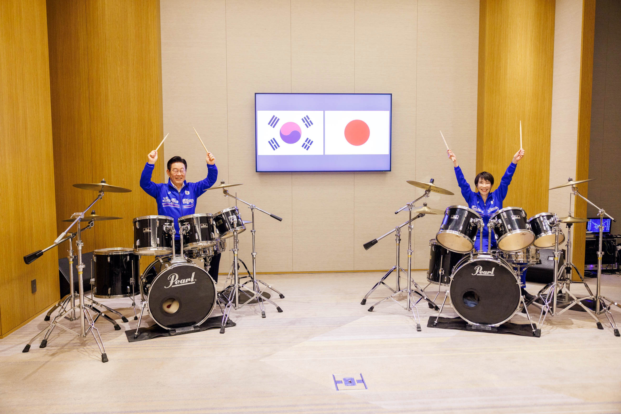 South Korean President Lee Jae-Myung and Japanese Prime Minister Sanae Takaichi play drums with their arms raised, in front of a screen displaying the South Korean and Japanese flags.
