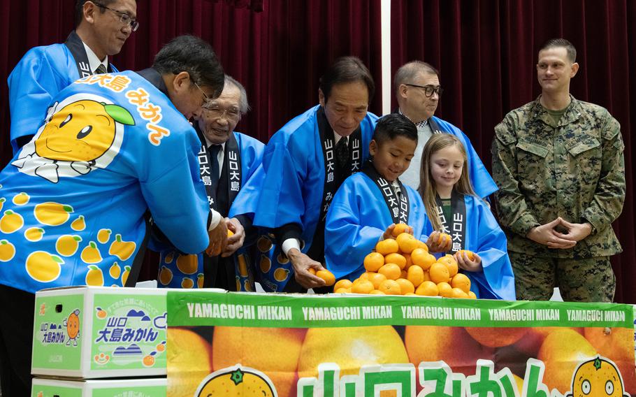 Two students in blue Japanese robes stand at a table laden with a pile of orange-colored mikan fruit, while Japanese men in blue robes and a military service member in camouflage uniform stand behind them.