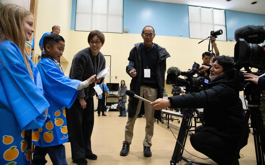 Two students in blue Japanese robes speak to a reporter who is kneeling beside a camera and holding a microphone in a school auditorium.