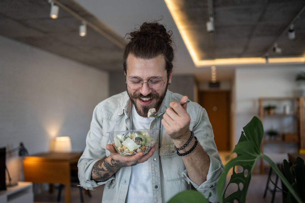 Young freelancer with glasses and tattoos savors a healthy salad during his work break in his modern, plant-filled home office, embracing a balanced lifestyle