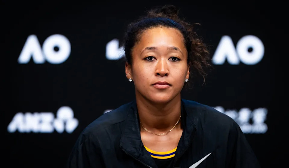 Naomi Osaka in a sporty outfit sits at a press conference table, looking forward