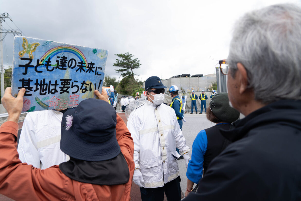 Manifestantes numa das entradas do lugar de construção de uma nova base militar na Baía de Henoko, em Okinawa. O cartaz em japonês diz: "O futuro dos nossos filhos não precisa de bases militares".
