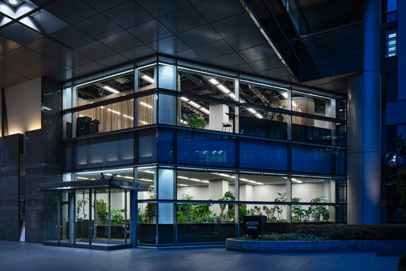 Modern glass building at night with two visible floors, illuminated interior lights, and plants inside; sign reading "NISSIN building" near the entrance.