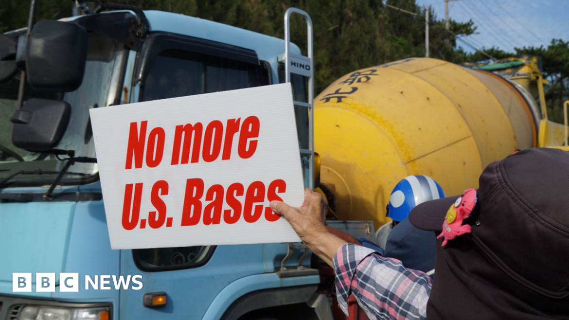 A man holds a white sign with red writing saying "no more US bases". He is wearing a plaid shirt and a cap, but his face cannot be seen. He stands in front of a blue and yellow concrete truck.
